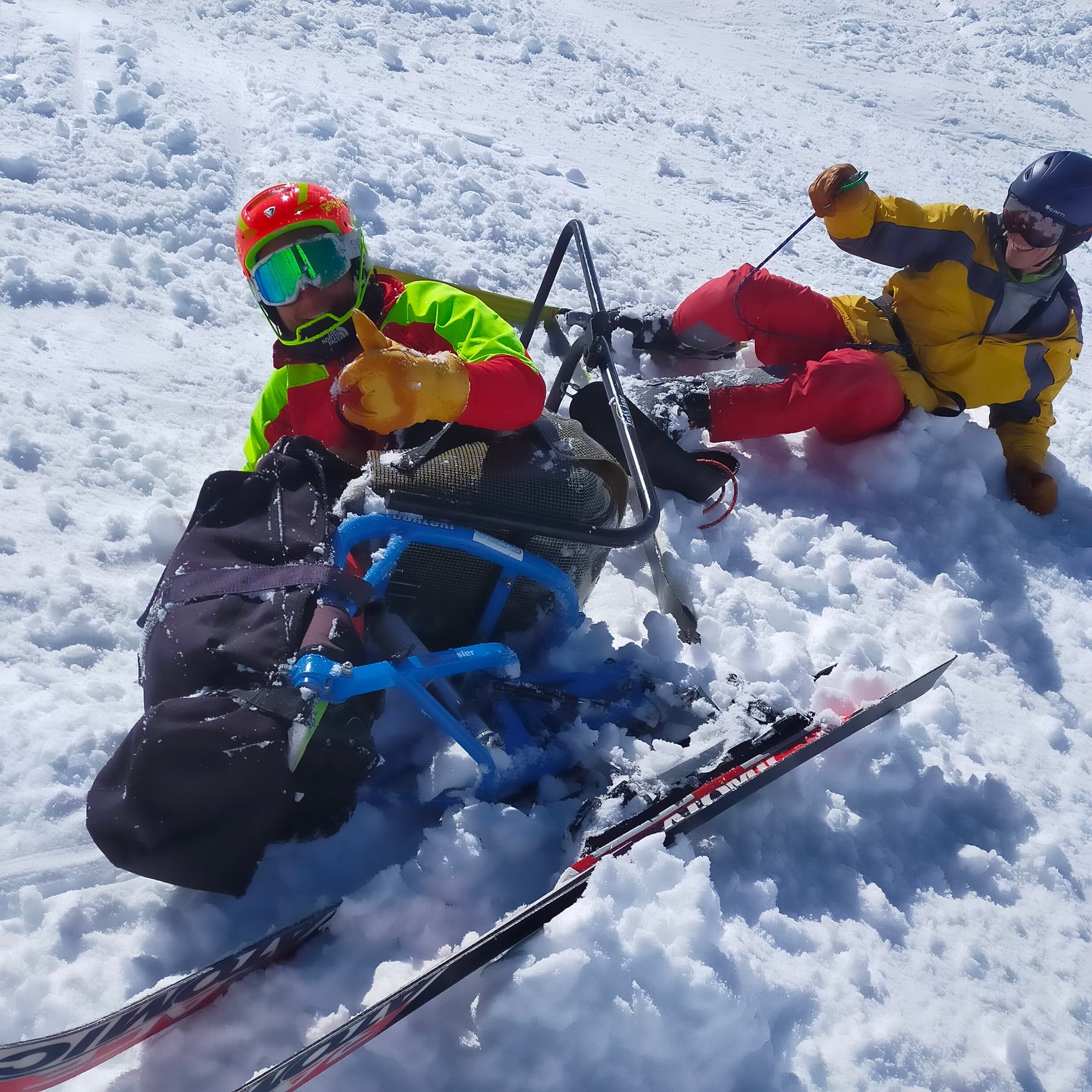 Deux sportifs dans la neige avec leur ski, la personne en premier plan lève le pouce en l'air, pris de côté. 
