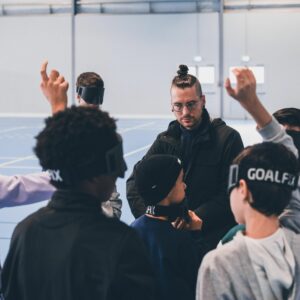 Une personne donne des consignes, plusieurs élèves en face d'elle lèvent la main, ils ont des masques sur leurs fronts pour bander leurs yeux.