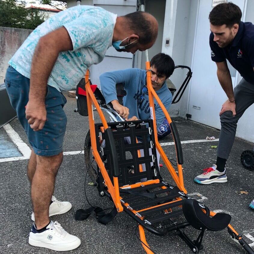 Trois personnes entrain de monter une joëlette pour être utilisée, pris de face. 