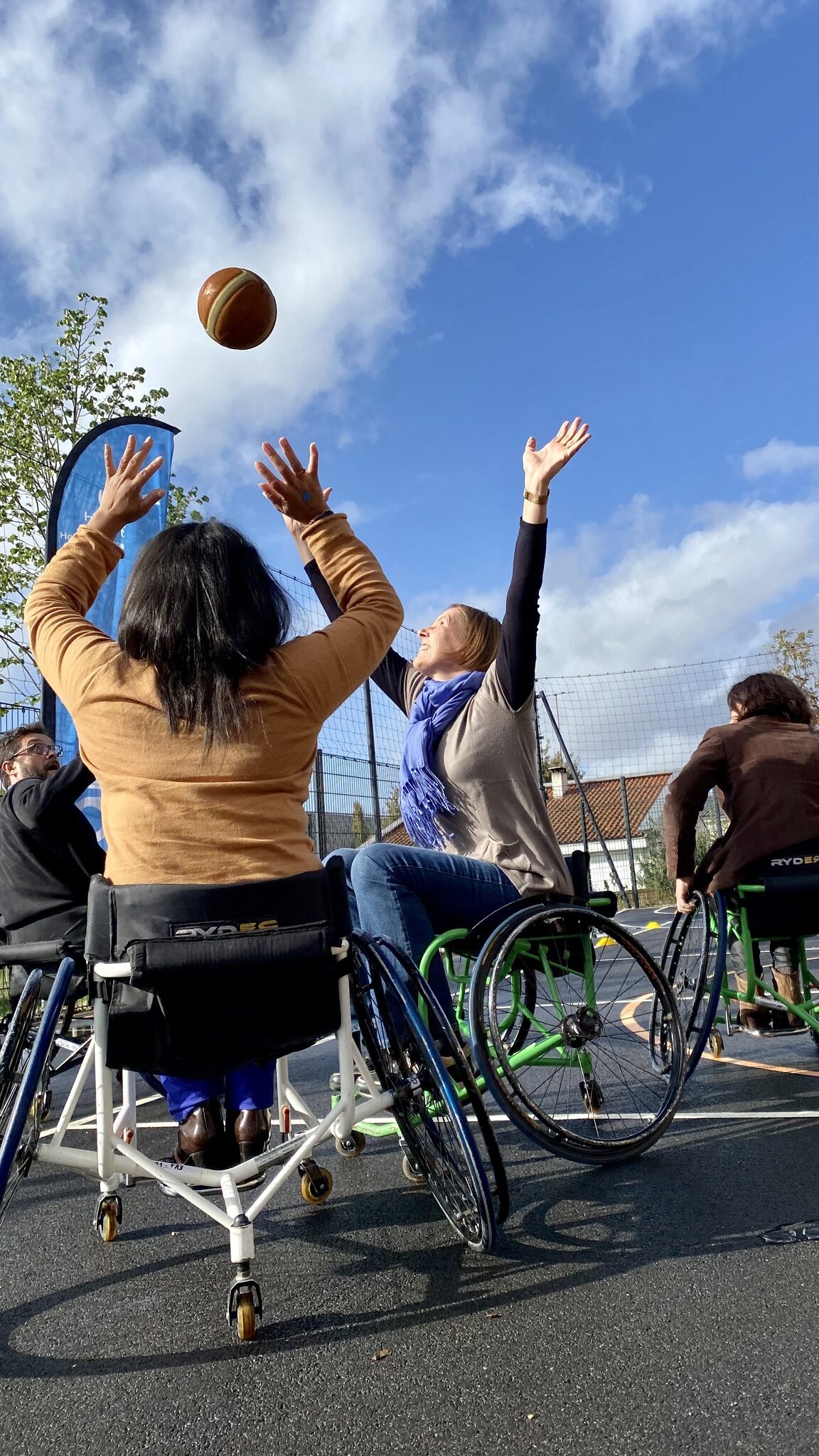 Quatre personnes pratiquant le basket-fauteuil en extérieur, le ballon est lancé en l'air, vue en contre-plongée.