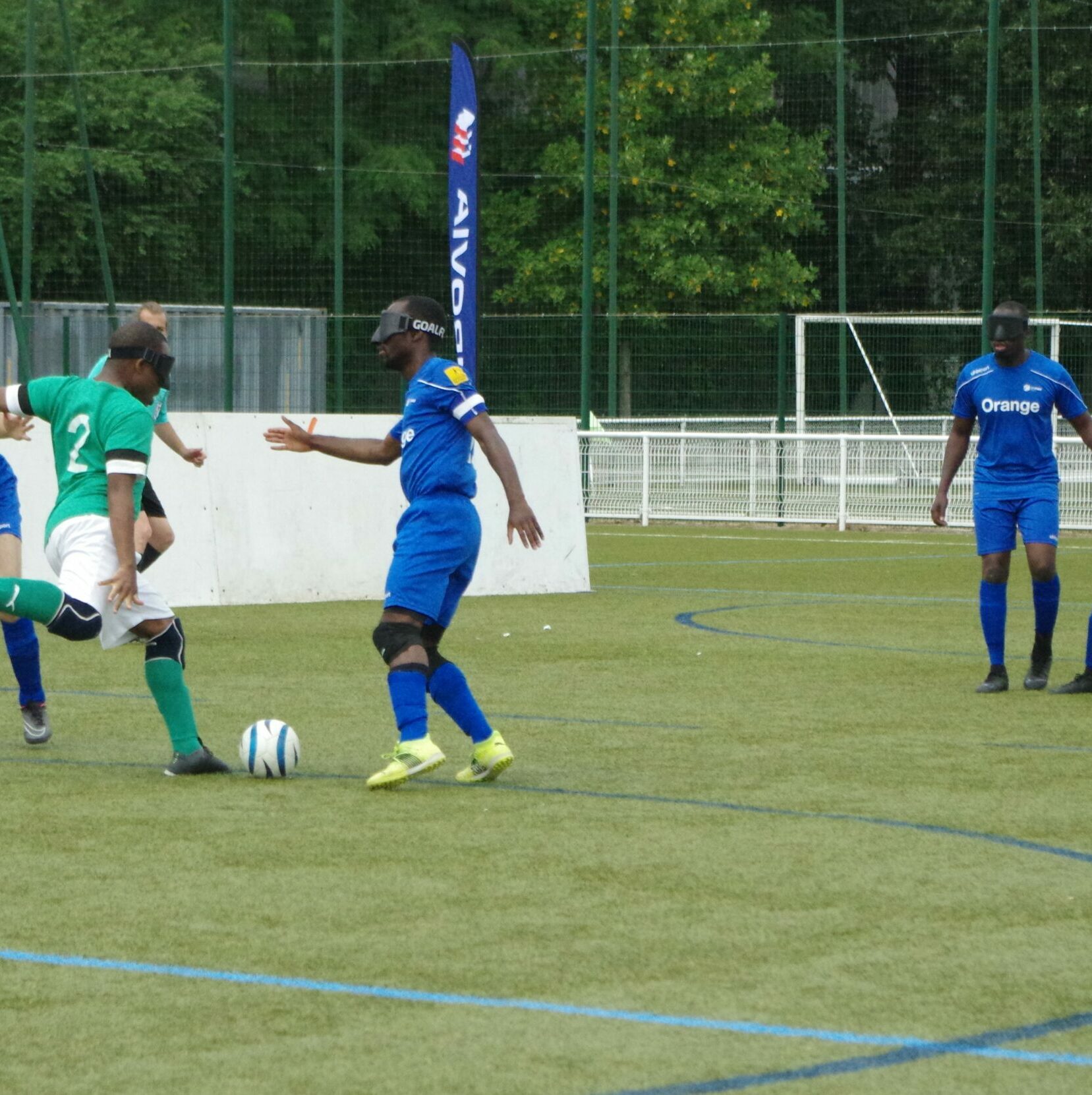 Plusieurs sportifs pratiquant le cécifoot, un sportif au maillot vert s'apprête à tirer alors que les adversaires en maillot bleu tentent de défendre. 