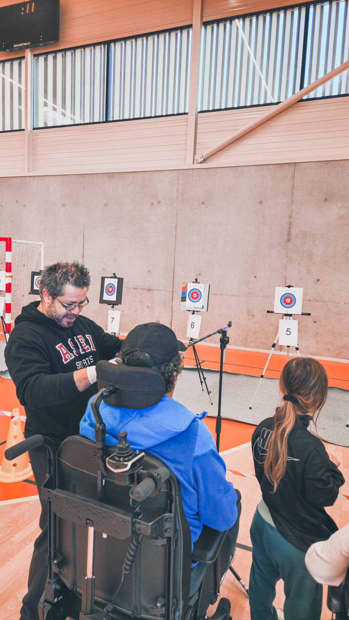 Un sportif et son accompagnateur en face des cibles, l'accompagnateur l'aide sur le matériel, sarbacane avec potence, pris de dos.