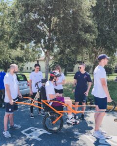 Une personne assise dans une joëlette tenue par une personne à l'avant et une personne à l'arrière, pris sur le côté